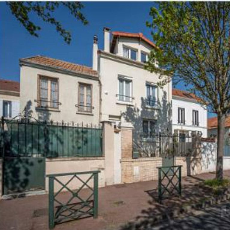 Maison blanche à deux étages avec balcons, entourée d'arbres et d'une clôture en fer.